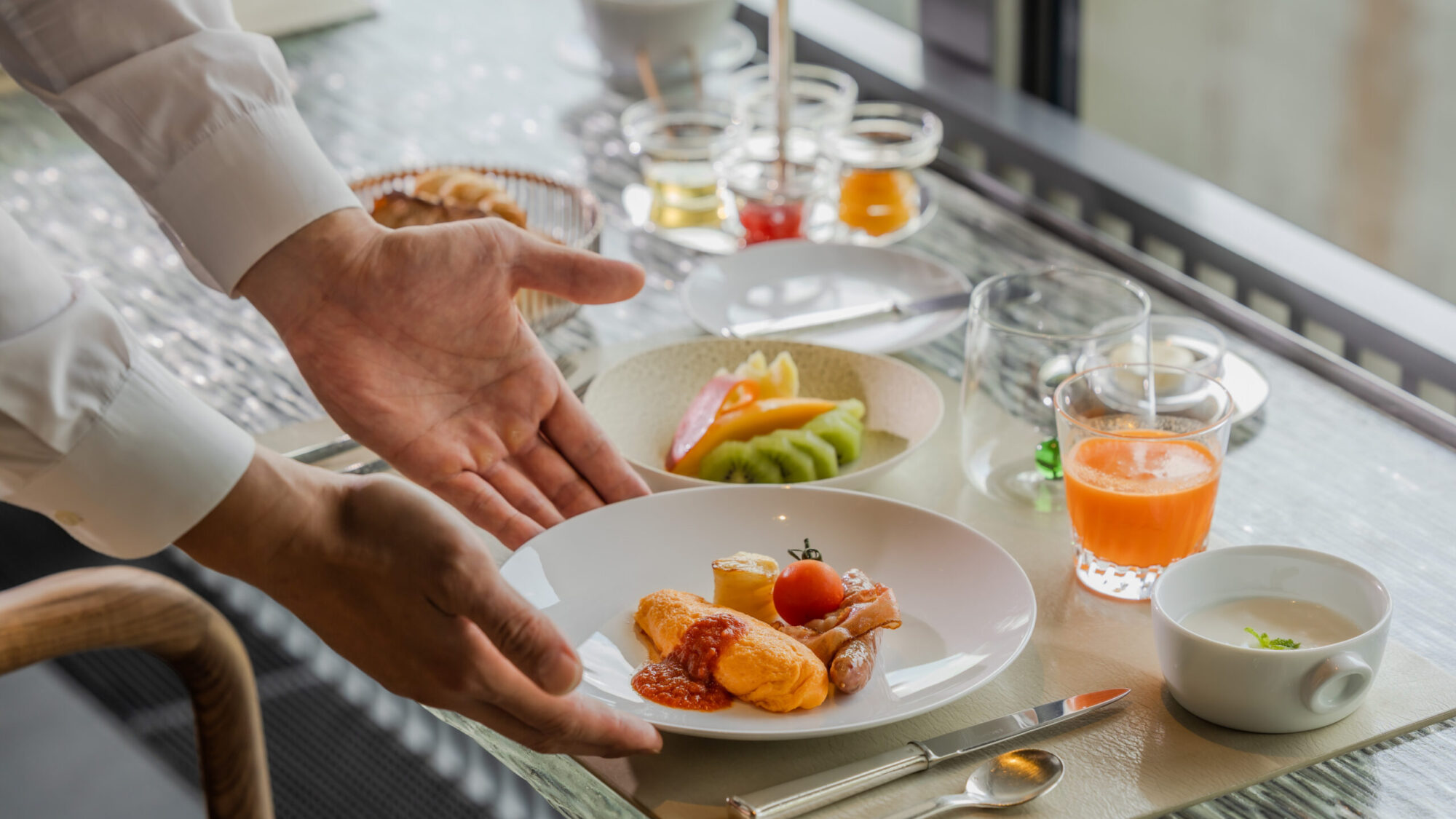 ホテルの朝食テーブルに並ぶ洋食セット。オムレツ、ソーセージ、スープ、フルーツ、ジュースなどが美しく配置され、スタッフが両手を広げて提供している様子。